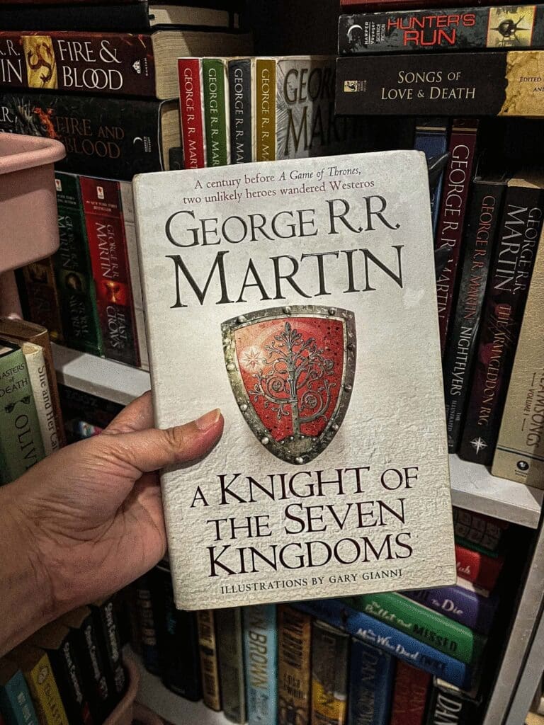 A hardcover copy of A Knight of the Seven Kingdoms in front of a collection of George R.R. Martin books in a shelf.